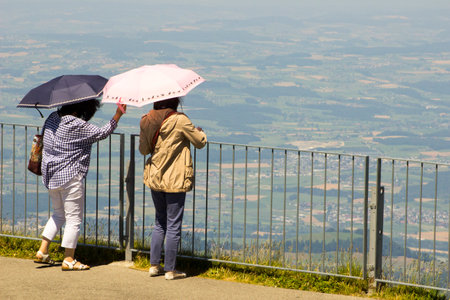 Two people standing side-by-side on a grassy hill, rain pouring down, both with umbrellas held high, prepared to face the wild weather togetherの写真素材