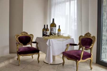 In this cozy dining room, a vibrant tablecloth drapes over a sturdy table, lined with chairs for sitting, while wine bottles and a vase of fresh flowers rest on the wall and light filters through the window, creating a warmの写真素材