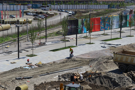 Workers are busy constructing a new urban pathway lined with trees and colorful murals. Heavy machinery and materials clutter the area, indicating ongoing development and landscaping efforts.の写真素材