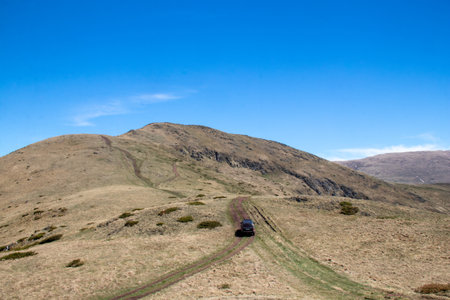 A vehicle navigates a rugged dirt path surrounded by expansive grassy hills. The landscape features a clear blue sky, enhancing the remote beauty of this mountainous area.の写真素材