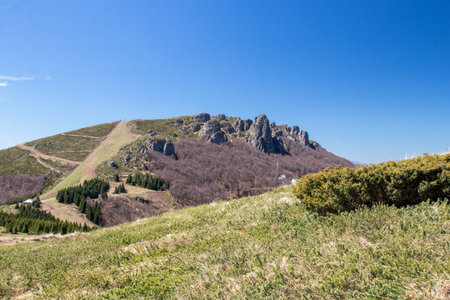 A stunning view of a mountain range features rocky outcrops and lush green fields under a clear blue sky. The scenery showcases the tranquility of nature in spring.の写真素材