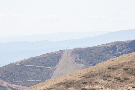 Rolling hills create a picturesque background for a distant ski lift, highlighting the serene beauty of the mountain landscape in the afternoon. The gentle slopes invite exploration.の写真素材