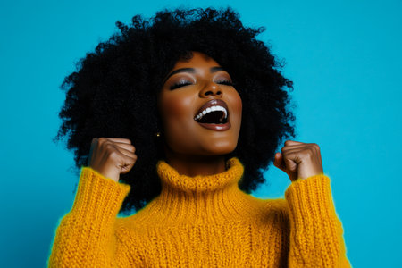 Excited dark skinned woman with afro hair clenching fists in victory against a blue backgroundの素材