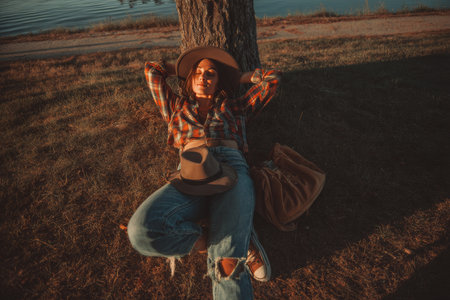 Woman relaxing on the ground with legs up, jeans and plaid shirt, by a lake in soft lightのeditorial素材