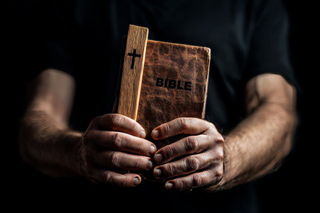 A man s hands holding a wooden cross and bible against a dark background in close up photographyの写真素材