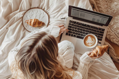 Freelancer working on laptop writes blog with coffee and croissant on bedsheet and tableの写真素材