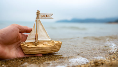 A person's hand gently cupping a miniature wooden sailboat with an "ideas" banner, set against a sunny shoreline with waves and soft sand.の写真素材