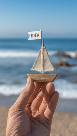A person's hand gently cradles a miniature wooden vessel with a sail and an "idea" flag, set against a sandy shoreline and vast ocean backdrop.の写真素材