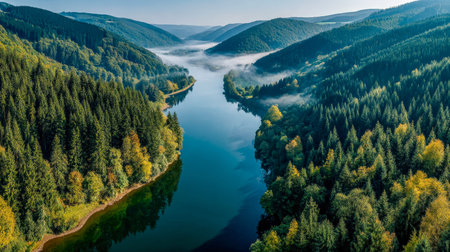 A tranquil waterway meanders through thick woodland hills shrouded in early morning mist, surrounded...