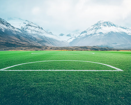 A lush green pitch stretches under a cloudy sky, with majestic snow-covered peaks forming a stunning backdrop for outdoor sports and recreation.の写真素材