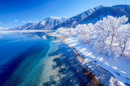 A pristine winter scene featuring towering snow-clad peaks, a glassy icy lake, and delicate frost-dusted trees beneath a vivid azure sky, evoking sereの写真素材