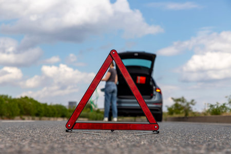 A warning triangle stands on the road with a car breakdown in the background, woman waiting for help...
