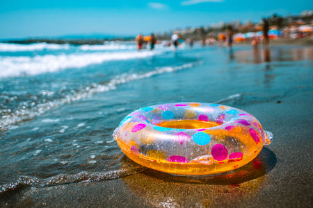 A vibrant, oversized float with rainbow hues lies partially deflated on the sandy edge, amidst crowded shorelines bustling with sunbathers and beachgoers enjoying seaside fun.の写真素材