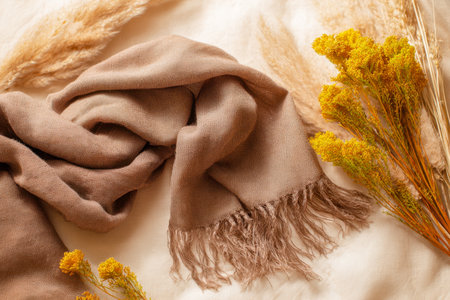 A warm brown woolen accessory with delicate fringes rests gracefully alongside bright yellow dried blooms, creating a charming autumn-inspired scene on a light fabric backdrop.の写真素材