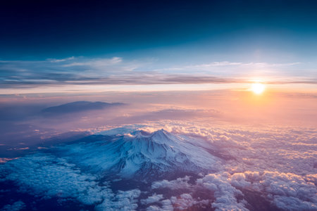 A birds-eye shot captures a rugged mountain summit blanketed in snow, glowing with dawn?s light above a sea of soft, swirling cloud layers.の写真素材
