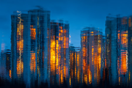 Soft, out-of-focus city lights shimmer on calm water as evening falls, creating a tranquil, dreamlike atmosphere with warm hues and blurred skyscraper silhouettes.のeditorial素材