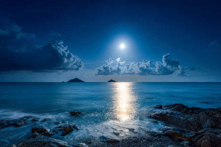 Serene nocturnal seascape with luminous moonlight shimmering across tranquil waters, dotted with distant islets and wispy clouds floating beneath a deep indigo sky.の写真素材