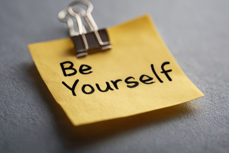 A bright yellow note, held by a sleek silver clip, features an inspiring quote amidst a calm gray backdrop, encouraging genuine self-expression and authenticity.の写真素材