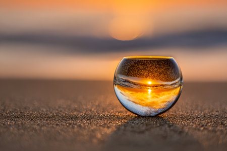 A close-up of a glass sphere resting on sand, capturing a vivid sunset and sky?s glow, creating a mesmerizing miniature world during warm evening light.の写真素材