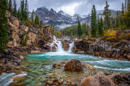 A tranquil turquoise stream cascades over rugged rocks amid lush pine woods, with towering snow-covered peaks and dreamy mist clouds enhancing autumn's peaceful charの写真素材