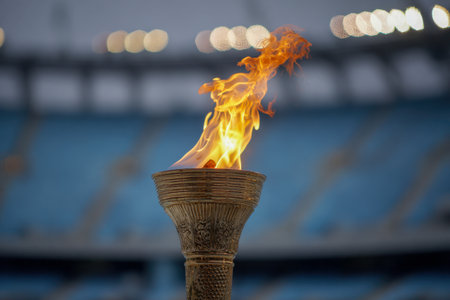 A vivid flame flickers fiercely from an intricately designed torch, set against a hazy stadium backdrop with glowing lights and out-of-focus spectators on a cloudy day.の写真素材