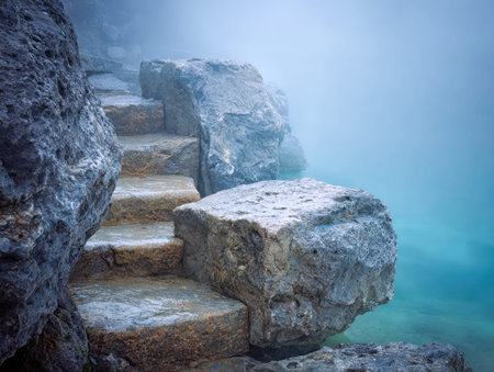 Weathered staircase hewn from cliffside stone rises gradually amid a gentle fog, overlooking tranquil teal waters in an untouched, peaceful wilderness.の写真素材