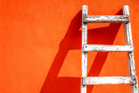 An aged wooden ladder leans against a boldly textured orange wall, its silhouette sharply defined by sunlight, evoking simple rustic elegance and quiet charm.の写真素材