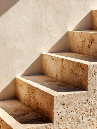 Warm golden sunlight highlights rough stone steps, creating crisp angular shadows that contrast beautifully with the sleek beige wall behind.の写真素材