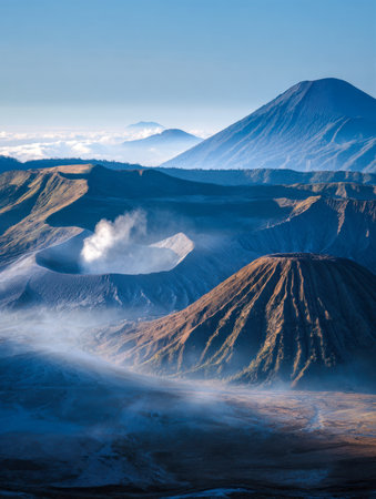 Dawn illuminates a rugged volcano, enveloped in swirling mist and releasing plumes of smoke, set beneath a crisp, azure morning sky.の写真素材