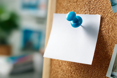 A minimalistic white card secured by a vibrant blue pin contrasts against a textured cork surface, with an out-of-focus office scene enhancing its purpose for notes or alの写真素材