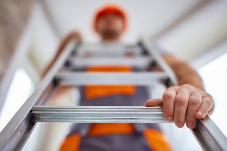 A diligent worker in a safety helmet ascends an aluminum ladder inside a building, with a close-up on hand gripping the rung during essential maintenance.の写真素材
