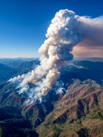 A towering, dense smoke cloud ascends over jagged forested peaks beneath a bright, cloudless sky, capturing the raw power and intensity of a fierce wildfire.の写真素材