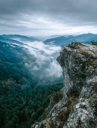 A rugged cliff offers a stunning vantage point over rolling, fog-shrouded valleys and distant mountain contours beneath a brooding, cloud-filled sky, showcasing wildの写真素材