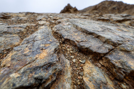 Rugged terrain featuring weathered stones, pronounced fissures, and loose gravel, set against a moody, cloud-filled sky amidst a remote mountain wilderness.の写真素材