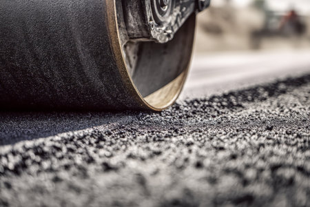 A powerful industrial machine flattens and presses newly laid road surface, its drum edge creating textured patterns on the gravel beneath bright sunlight.の写真素材