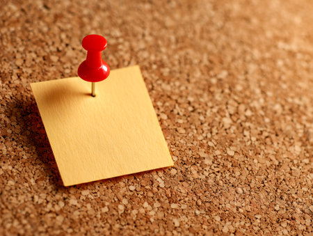 A vibrant yellow memo secured with a bold red pin on a rough cork surface, offering space for personalized notes or important highlights within a busy workspace.の写真素材