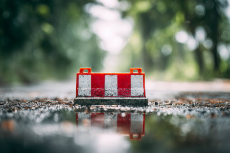 A tiny striped barricade pauses on glistening pavement, its vivid pattern mirrored perfectly in a surrounding puddle, with a soft-focus greenery scene behind.の写真素材