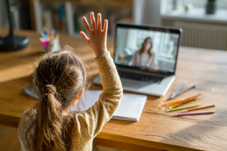 A cheerful girl eagerly participates in a virtual class from her cozy home, surrounded by colorful pencils and notebooks on a wooden desk beside her laptop.の写真素材