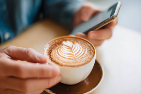 A calm moment captured as a person leisurely browses on their smartphone, cradling a beautifully crafted cappuccino on a warm wooden surface bathed in gentle naturalの写真素材