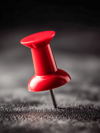 A bold crimson thumbtack strikes a textured dark background, illuminated by intense lighting that...