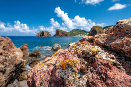 Bold, multicolored stones with rich textures dominate the foreground, while gentle ocean swells fade into a verdant hillside under a clear, cloud-dappled sky.の写真素材