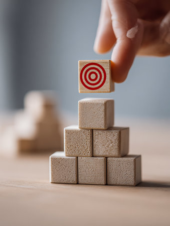 A hand carefully places a marked wooden block atop a rising tower of plain blocks, illustrating strategic planning and the pursuit of objectives.の写真素材