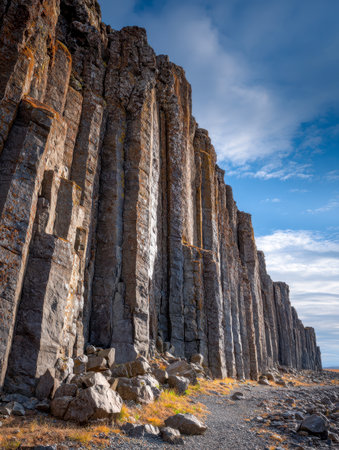 Towering black volcanic pillars rise sharply against a bright blue sky, casting shadows on rugged terrain as hikers traverse a scenic rocky trail on a sunny day.の写真素材