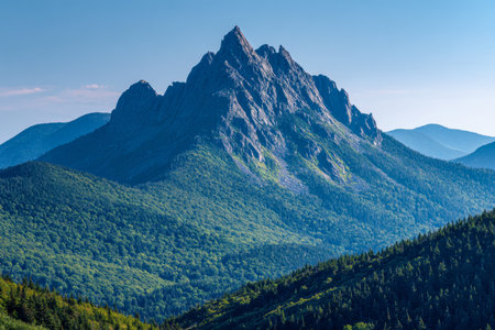 Towering rugged summit rises above vibrant green hills, framed by dense forests under a bright azure sky, evoking tranquility in untouched wilderness.の写真素材