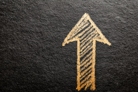 A vivid yellow chalk arrow pointing upward on a dark, textured background, embodying momentum,...
