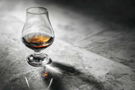 A slender glass holds a splash of rich amber spirit, creating an elongated shadow on rough stone under gentle, subdued illumination.の写真素材