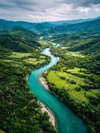 An expansive shot from above captures a vibrant turquoise stream meandering through dense green woodlands and expansive meadows, set beneath a striking, stormy sky iの写真素材