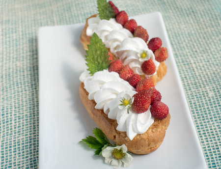 Fresh cream eclairs with whipped cream and strawberries on white plate and cup of coffee on white...
