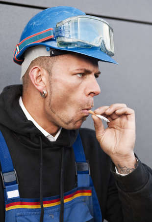 Manual worker in protectice clothes and in a blue hard hat smoking outdoorsの写真素材