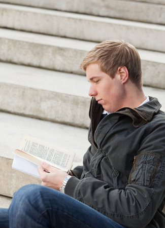 Young blond handsome chap reading book while sitting on the stairs outdoors
